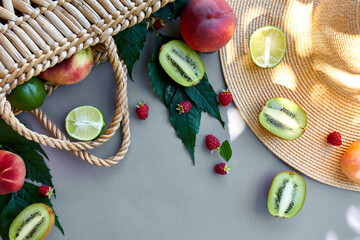 Flat lay of Straw Hat And Bag With Fruits raspberry, peach, kiwi, lime  on grey background in sunlight, summer time. Grocery concept, copy space, top view.