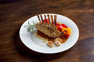 Appetizing roasted rack of lamb in nuts, with sauce and grilled vegetables, served in a white plate on a wooden background.