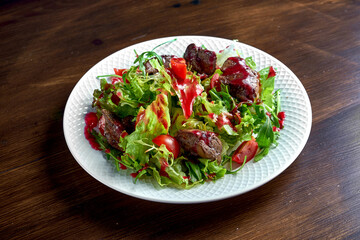 Healthy and dietary salad mix with berry sauce, fried chicken liver and cherry tomatoes, served in a white plate on a wooden background
