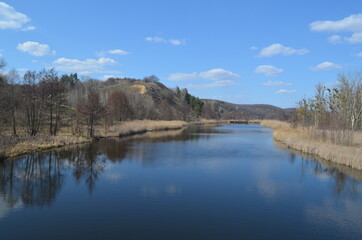 Beautiful landscape with a wide blue river and high hill on the horizon. Reflection of the sky and clouds in the water. Early spring nature in sunny warm weather.