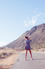 Naklejka premium young woman with long brunette hair posing wearing a hat and flower dress walking along a path in a dry and hilly yellow spot in the late afternoon