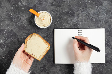 Female hands write notes in a notebook and hold a sandwich with hummus on a dark background. Healthy snack during work and study