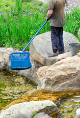 A utility worker cleans a pond of mud with a long net in a city park.