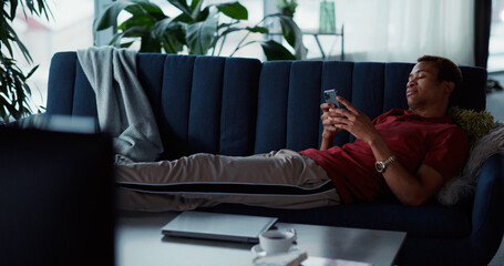Casual afro-american young man resting down on couch using smartphone playing video game networking surfing internet news. Candid lifestyle. Stay home.