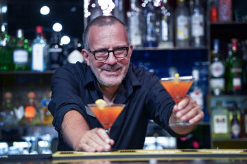 Elderly bartender with eyeglasses serving cocktail at the bar. Evening time