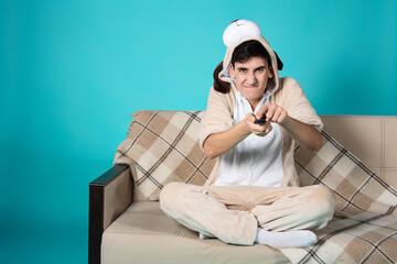 A funny guy in pajamas sits in front of the TV with a remote control. Blue background. Beige sofa.