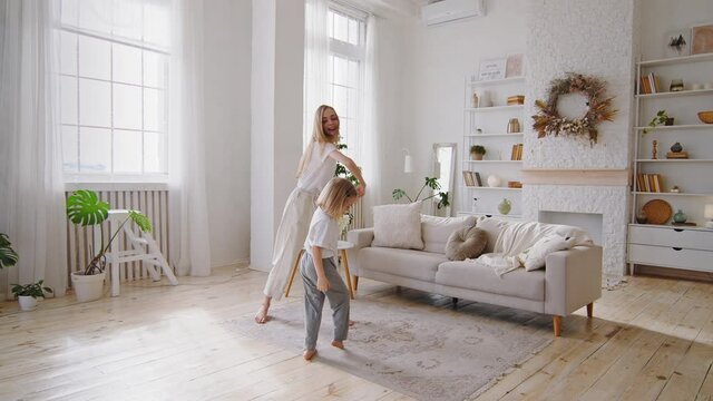 Young blonde mother mom dancing with little daughter small girl at home living room interior, cheerful mum and active child kid family enjoy dance have fun moving to music together at weekend indoor