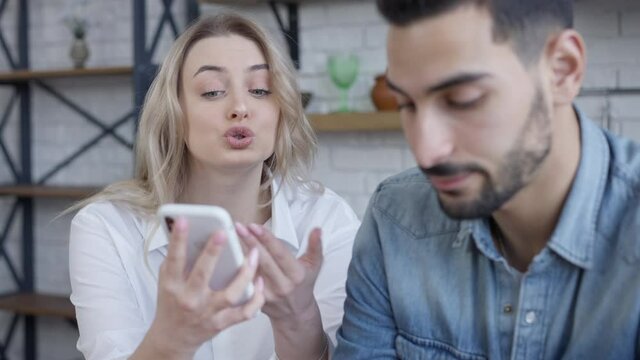 Serious slim beautiful woman talking with blurred man gesturing and pointing at smartphone. Portrait of dissatisfied Caucasian girlfriend arguing with cheating boyfriend at home indoors