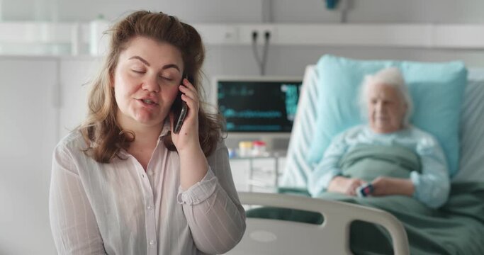 Woman in hospital ward talking on smartphone with sick aged mother in hospital bed on background