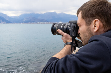 Casual photographer taking photos of the sea and mountains. Brutal handsome young man using his big camera during traveling. Professional photographer journalist catching the frames outside.