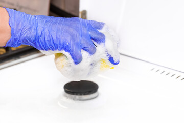 The hand of a housewife in a rubber household glove holds a sponge with foam over the gas stove and is engaged in home cleaning kitchen, close-up