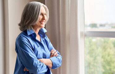 Mature happy laughing 60s middle aged business woman standing indoors in home office, arms crossed. Senior elder elegant serene woman looking at window thinking of positive vision enjoying wellbeing.