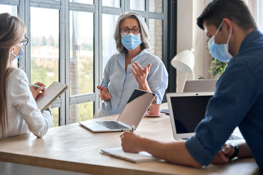 Senior Older Educator Working With Computers In Office With Team Wearing Medical Masks During Covid Lockdown.Adult Experienced Business Woman Is Holding Meeting With Female Assistant And Male Manager.