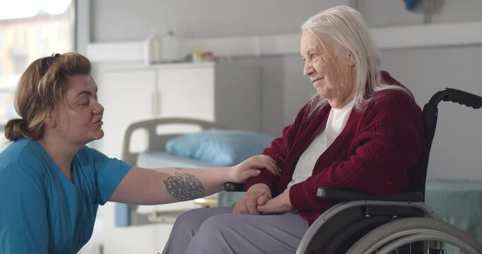 Friendly staff caregiver of nursing home talking to senior woman in ward