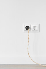 New white plastic European electrical outlet isolated on white plastered wall. An electrical plug with a vintage cable is plugged into an outlet. Gray wood floor. Copy space