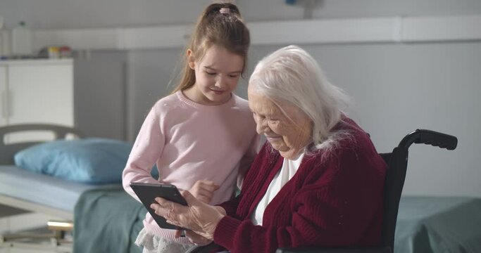 Little girl hugging disabled grandmother in wheelchair using digital tablet in nursing home