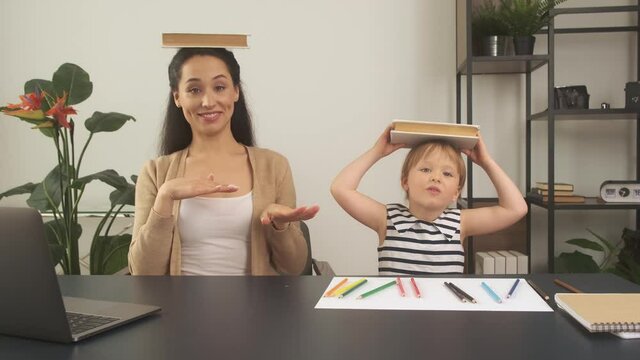 Mother and daughter having fun during remote learning at home, sitting in yoga pose with books on a head, isolation concept