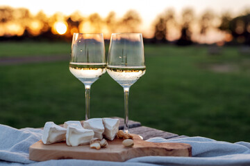 Two glasses of white wine and a wooden plate with cheese and nuts served outside at sunset. - obrazy, fototapety, plakaty