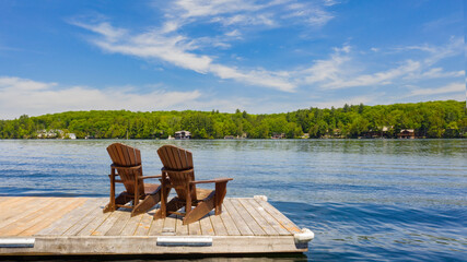 Two Adirondack chairs on a wooden dock facing a lake in Muskoka, Ontario Canada during a sunny...