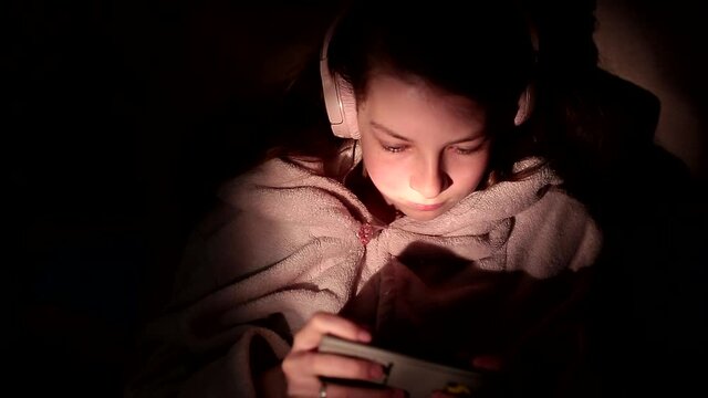 a teenage girl in wireless headphones at night in bed with a smartphone in her hands listens to streaming music