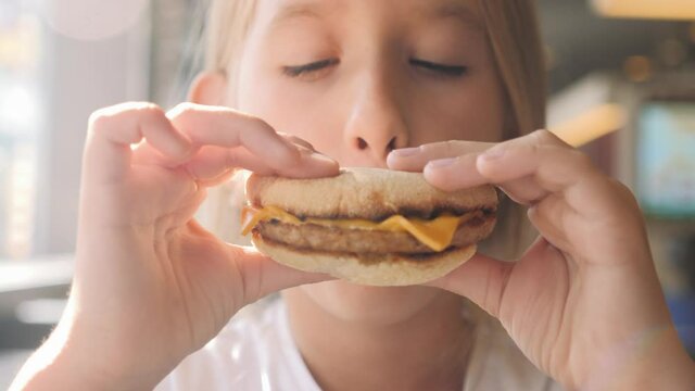 Fast food concept. Young girl in a cafe eats a hamburger. Child who takes a bite and chaos a piece of hamburger with gusto. Unhealthy eating. Hungry kid eating junk food.