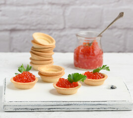 red caviar in a glass jar and round tartlets on a white table