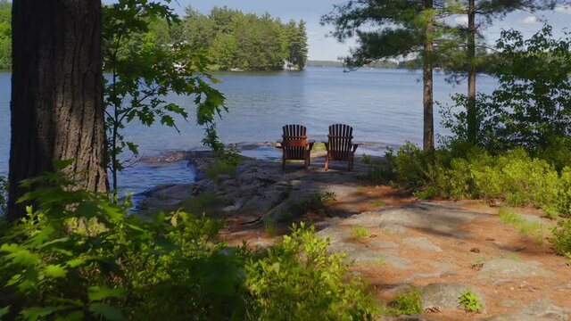 Drone flying over two Adirondack chairs sitting on a rock formation facing the blue waters of a lake in Ontario, Canada