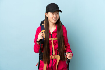 Young Chinese girl with backpack and trekking poles over isolated blue background looking up while smiling