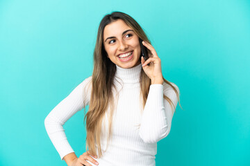 Young caucasian woman using mobile phone isolated on blue background posing with arms at hip and smiling