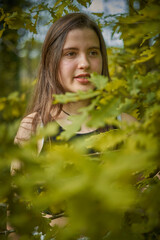 Naklejka premium Light-skinned young woman with long hair, black top and necklace among the leaves and branches of an oak tree. girl in nature. teenage girl among the branches of a green oak tree smiling and pensive.
