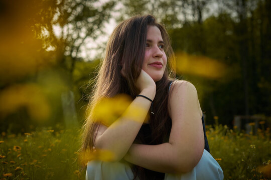 Light-skinned young woman with long hair, black top and white pants sitting in field of yellow flowers smiling and thinking. girl in nature. teenager with daisies thinking and smiling. eco person
