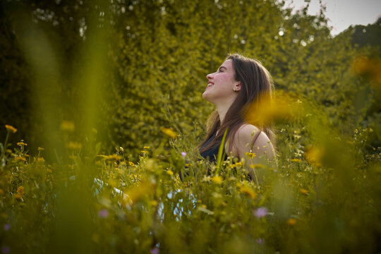 Light-skinned young woman with long hair, black top and white pants sitting in field of yellow flowers smiling and thinking. girl in nature. teenager with daisies thinking and smiling. eco person