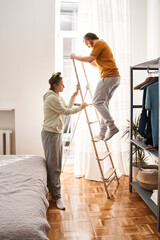Fototapeta na wymiar Woman holding ladder while her man standing on it during the hanging curtains