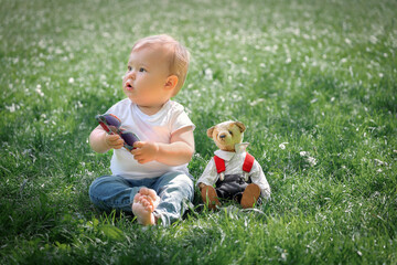 little boy 1 year old sitting on green grass in park with teddy bear toy, happy childhood