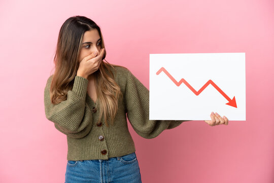 Young caucasian woman isolated on pink background holding a sign with a decreasing statistics arrow symbol with surprised expression