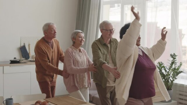 Slow motion medium long of happy multiethnic senior friends dancing conga at home around dining table