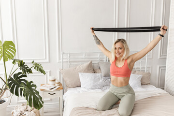 Fototapeta na wymiar Happy young woman training in the morning, sitting on bed, doing exercises for the arms. Sportive blonde-hair girl stretching out, working out at home. Healthy lifestyle concept