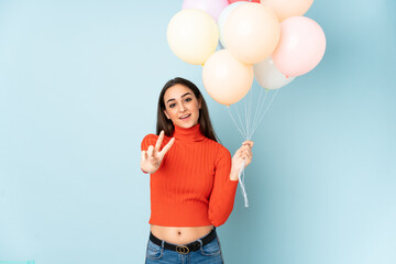 Young woman catching many balloons isolated on blue background smiling and showing victory sign