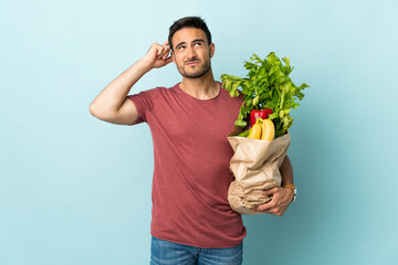 Young caucasian man buying some vegetables isolated on blue background having doubts and with...