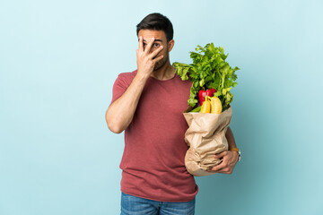 Young caucasian man buying some vegetables isolated on blue background covering eyes and looking...