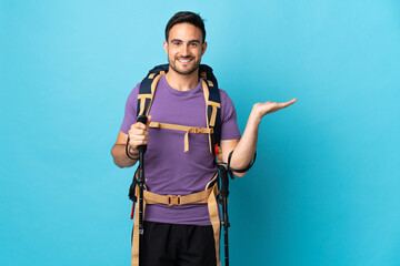 Young caucasian man with backpack and trekking poles isolated on blue background holding copyspace imaginary on the palm to insert an ad