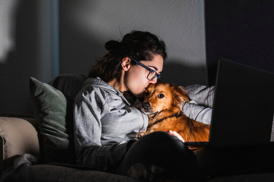 A young woman kisses her dog while watching a movie on her computer at night. At home concept. Animal love.