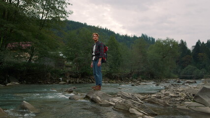 Tourist standing on rocky shore of river. Joyful man looking green landscape