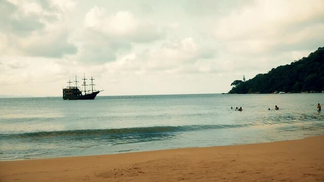 A medieval ship is getting closer to the beach to dock on the port on a cloudy day with people on the water. Old ship similar to Columbus vessel, exploring america with navigator skills on calm sea