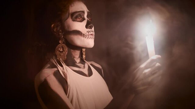 Hispanic woman makeup as a Catrina blowing smoke and holding a candle during a funeral in Mexico. Halloween costume.  Slow-motion