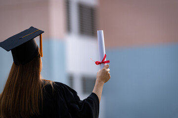 A young happy Asian woman university graduate in graduation gown and mortarboard holds a degree certificate celebrates education achievement in the university campus.  Education stock photo - obrazy, fototapety, plakaty