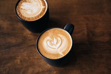 Two cappuccino coffee with latte art on wooden table. Black cups and beautiful milk foam. Soft focus