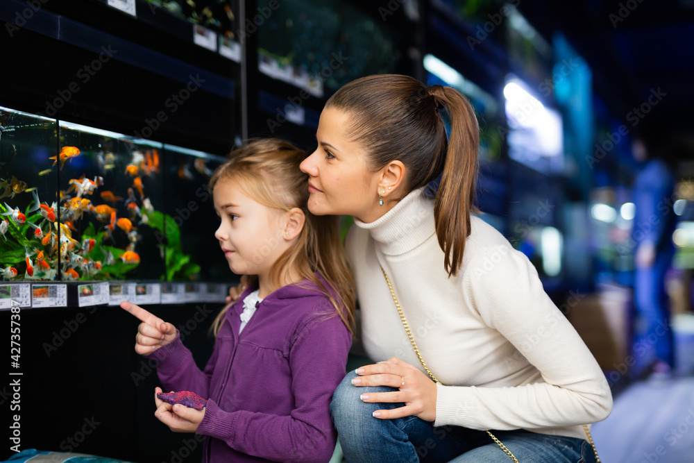 Wall mural Interested cute 5 year old girl with her mother looking at colorful tropical fish in aquariums in pet shop..