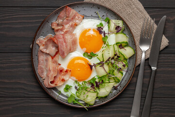 Continental breakfast - eggs, bacon, micro greens and cucumber slices on a plate on the table. Healthy breakfast. Top view