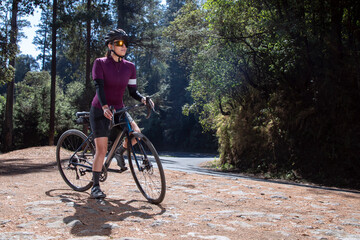 Fototapeta premium Joven ciclista descansando con su bicicleta al lado de la carretera , usando casco y lentes del sol, en medio del bosque. deporte al aire libre